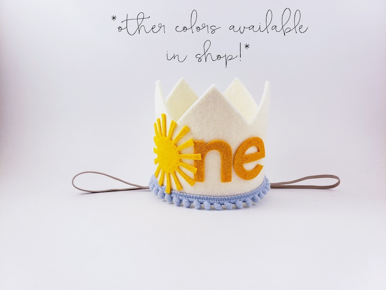 May include: A cream felt crown featuring a yellow sun design and the word "one" in gold. The crown is trimmed with light blue pom-poms and has a grey elastic band. Text in the image says "other colours available in shop!"
