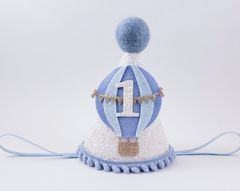 Könnte beinhalten: Eine weiße und blaue Filz-Partymütze mit einem blauen und weißen Heißluftballon-Design. Der Ballon hat eine goldene Glitzerzahl "1" und ein goldenes Glitzerband. Die Mütze hat einen blauen Filz-Kinngurt und einen weißen Pompon-Besatz.
