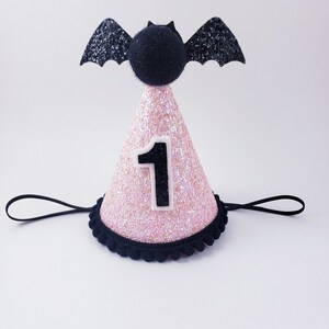 May include: Pink glitter party hat with a black bat on top and a white number one on the front. The hat has a black elastic band.