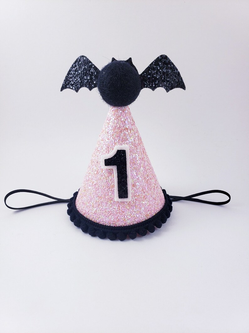 May include: Pink glitter party hat with a black bat on top and a white number one on the front. The hat has a black elastic band.