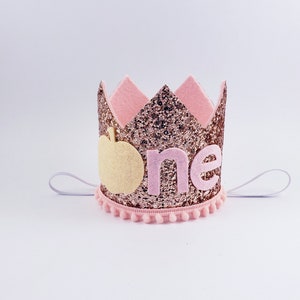 May include: Pink and gold glitter crown with a white felt apple and the word "one" in pink felt letters. The crown has a white elastic band.