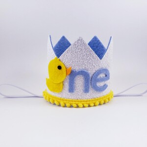 May include: A white felt crown with a blue felt trim and the word "one" in blue felt letters. A yellow felt duck is on the side of the crown. The crown has a white elastic band.