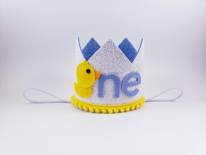 May include: A white felt crown with a blue felt trim and the word "one" in blue felt letters. A yellow felt duck is on the side of the crown. The crown has a white elastic band.