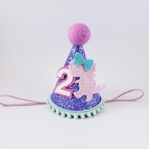 May include: Purple glitter party hat with a pink dinosaur, a blue bow, and the number "2" on the front. The hat has a light pink elastic band.