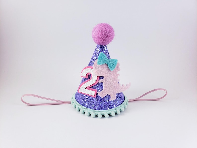 May include: Purple glitter party hat with a pink dinosaur, a blue bow, and the number "2" on the front. The hat has a light pink elastic band.