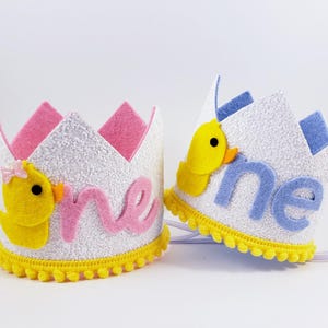 May include: Two white felt crowns with yellow pom-poms around the base. One crown has a pink felt "one" and a yellow felt duck with a pink bow. The other crown has a blue felt "one" and a yellow felt duck.