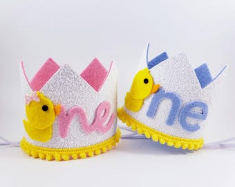 May include: Two white felt crowns with yellow pom-poms around the base. One crown has a pink felt "one" and a yellow felt duck with a pink bow. The other crown has a blue felt "one" and a yellow felt duck.