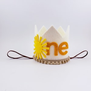 May include: A white felt crown featuring a yellow sun and the word "one" in orange. It has a brown string and a tan trim with small pom-poms. This crown is ideal for a first birthday celebration.