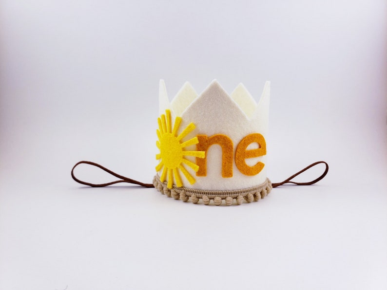 May include: A white felt crown featuring a yellow sun and the word "one" in orange. It has a brown string and a tan trim with small pom-poms. This crown is ideal for a first birthday celebration.