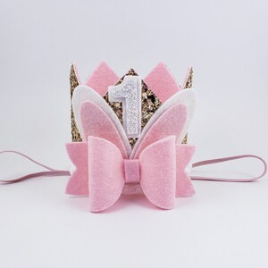 May include: Pink felt crown with a gold glitter number one and white felt bunny ears. The crown is attached to a pink elastic headband.