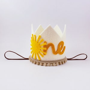 May include: A cream felt crown featuring a yellow sun and the word "one" in gold. The crown has a brown elastic band and a tan pom-pom trim. Ideal for a first birthday, this crown is a charming accessory.