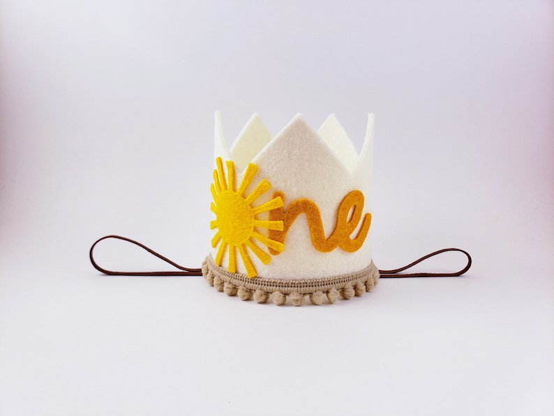 May include: A cream felt crown featuring a yellow sun and the word "one" in gold. The crown has a brown elastic band and a tan pom-pom trim. Ideal for a first birthday, this crown is a charming accessory.