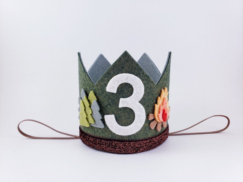 May include: A green felt crown with a white felt number 3, a felt campfire, and felt pine trees. The crown has a brown felt band.