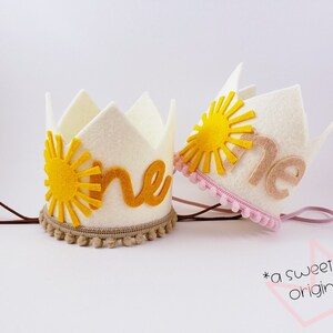 May include: Two white felt crowns, each with a yellow sun design and the word "one". One crown has a brown trim with pom-poms, the other a pink trim. Both have elastic bands. These are birthday crowns. The text "a sweetcakes original" is also visible.
