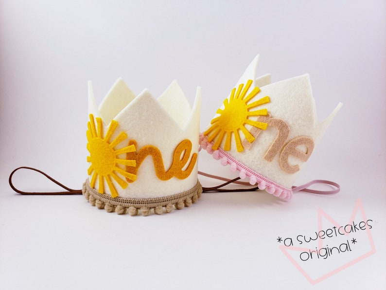 May include: Two white felt crowns, each with a yellow sun design and the word "one". One crown has a brown trim with pom-poms, the other a pink trim. Both have elastic bands. These are birthday crowns. The text "a sweetcakes original" is also visible.