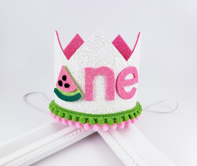 May include: White felt crown with pink felt trim and a pink felt "one" with a green and red felt watermelon slice on the side. The crown has a green and pink pom-pom trim.