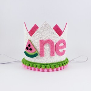 May include: White felt crown with pink felt trim and a watermelon applique. The crown has a green and pink pom-pom trim and says "one" in pink felt letters.