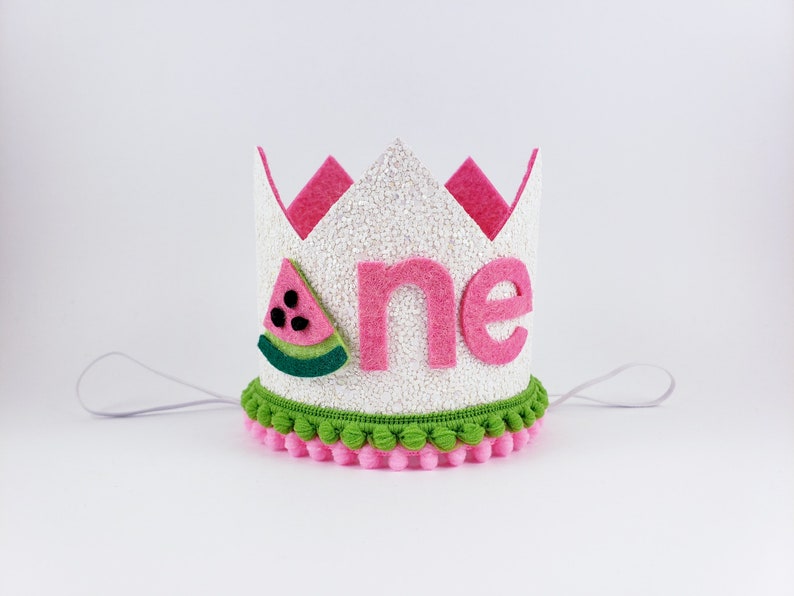 May include: White felt crown with pink felt trim and a watermelon applique. The crown has a green and pink pom-pom trim and says "one" in pink felt letters.