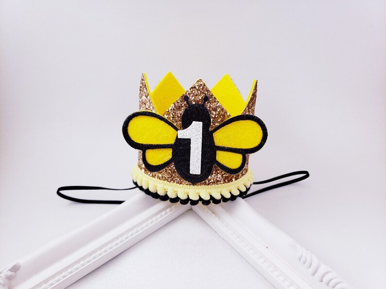 May include: Gold glitter bee crown with a yellow felt bee and the number 1 in white. The crown has a black elastic band.