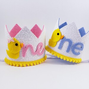 May include: Two white felt crowns with yellow pom-poms around the base. One crown has a pink felt "one" and a yellow felt duck with a pink bow. The other crown has a blue felt "one" and a yellow felt duck. Both crowns have white felt and glitter.