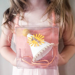 May include: A white felt party hat with a yellow sun and gold glitter number one. The hat is in a clear plastic box with a white ribbon.