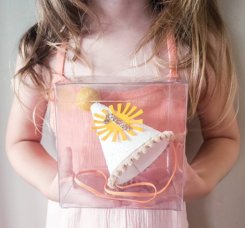 May include: A white felt party hat with a yellow sun and gold glitter number one. The hat is in a clear plastic box with a white ribbon.