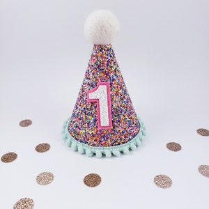 May include: A colorful cone-shaped party hat with a white pom-pom on top. The hat is covered in rainbow glitter and has a pink felt number "1" on the front. The hat is surrounded by gold glitter confetti.