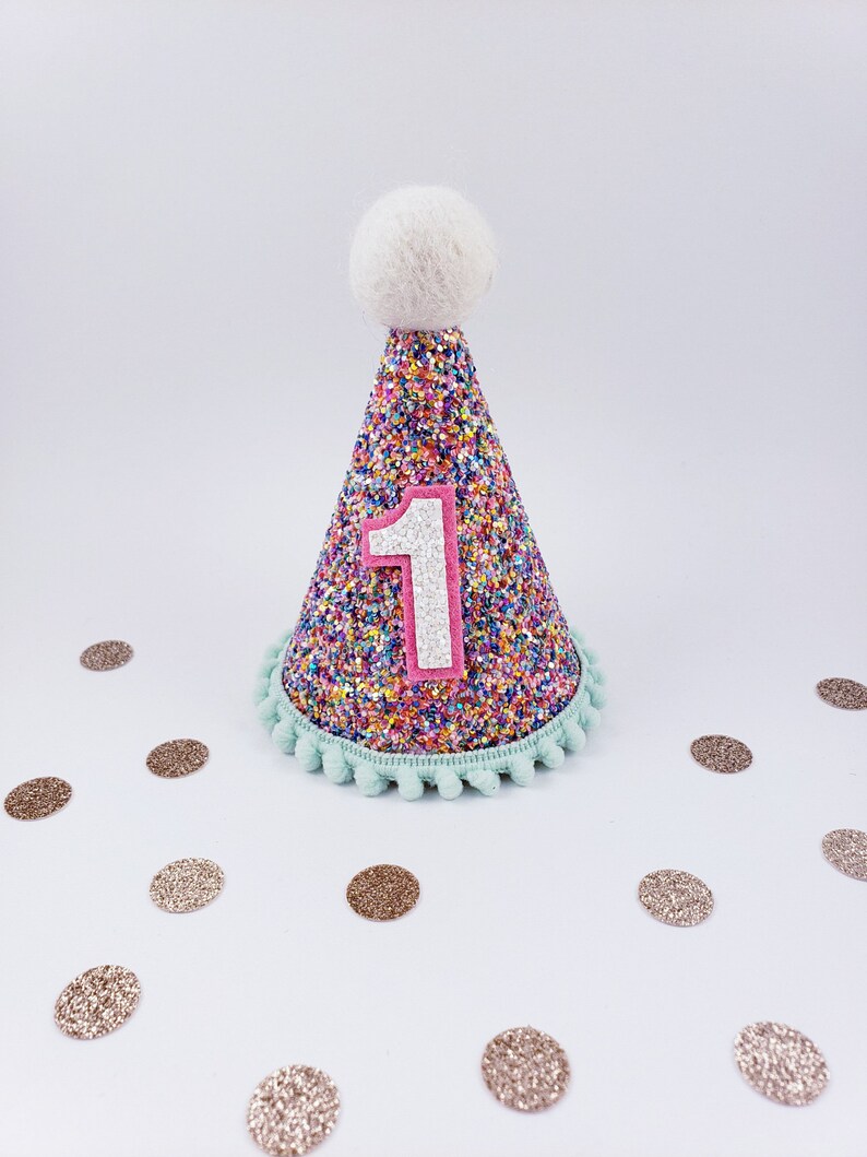 May include: A colorful cone-shaped party hat with a white pom-pom on top. The hat is covered in rainbow glitter and has a pink felt number "1" on the front. The hat is surrounded by gold glitter confetti.