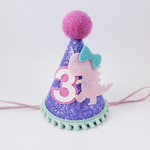 May include: Purple glitter party hat with a pink felt dinosaur, a blue bow, and a white felt number 3. The hat has a pink elastic band and a green pom-pom trim.