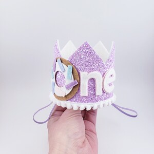 May include: Purple glitter crown with white felt trim and a donut design. The crown says "one" in white felt letters.