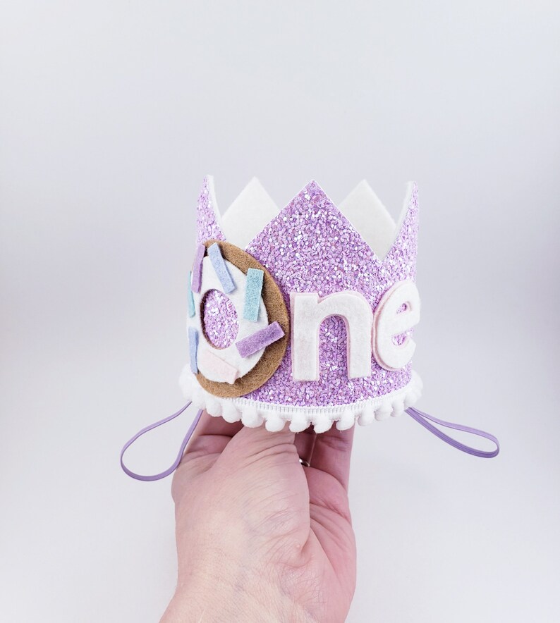 May include: Purple glitter crown with white felt trim and a donut design. The crown says "one" in white felt letters.