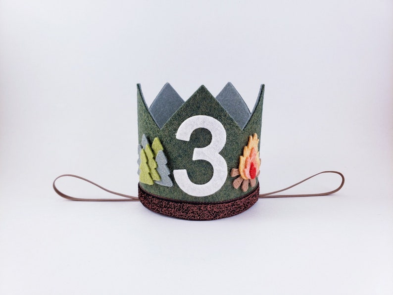 May include: Green felt crown with a white felt number 3, a felt campfire with orange flames, and green felt trees. The crown has a brown felt band with glitter and a brown felt tie.