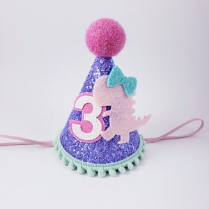 May include: Purple glitter party hat with a pink felt dinosaur, a blue bow, and the number 3 on the front. The hat has a green pom-pom trim and a pink felt ball on top.
