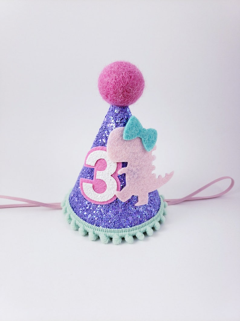 May include: Purple glitter party hat with a pink felt dinosaur, a blue bow, and the number 3 on the front. The hat has a green pom-pom trim and a pink felt ball on top.