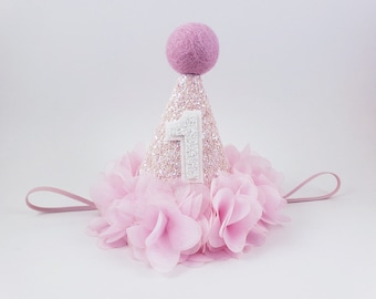 May include: Pink glitter party hat with a white felt number one and a pink tulle trim. The hat has a pink felt pom pom on top and a pink elastic band.
