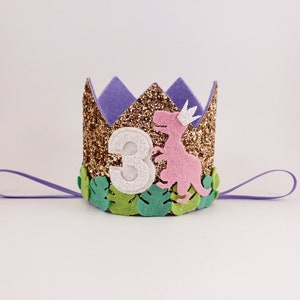 May include: A gold glitter crown with a purple felt top and green felt leaves. A pink felt dinosaur with a white crown is on the front of the crown. The number "3" is on the front of the crown in white felt.