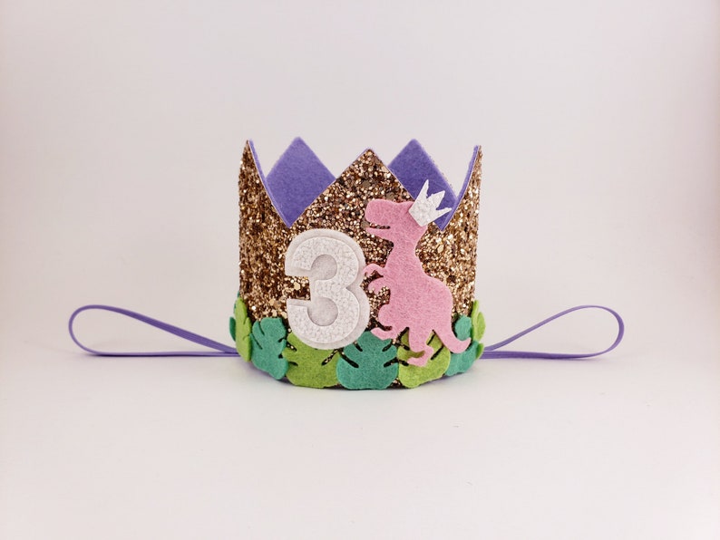 May include: A gold glitter crown with a purple felt top and green felt leaves. A pink felt dinosaur with a white crown is on the front of the crown. The number "3" is on the front of the crown in white felt.