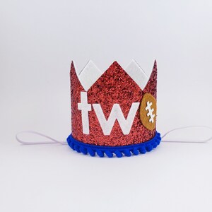 May include: Red glitter crown with white felt 'two' and a brown felt football with a white stitch design. The crown has a blue pom-pom trim and a white elastic band.