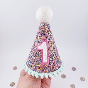 May include: A pink and white glitter party hat with a white pom-pom on top and a white number "1" on the front. The hat has a light blue trim with pom-poms.