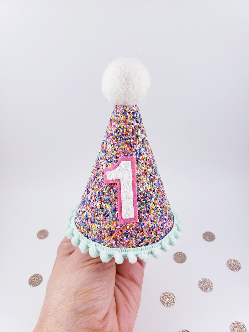 May include: A pink and white glitter party hat with a white pom-pom on top and a white number "1" on the front. The hat has a light blue trim with pom-poms.