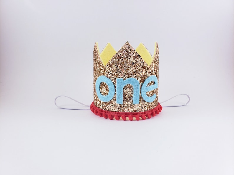 May include: Gold glitter crown with a blue felt "one" on the front. The crown has a red pom-pom trim and a white elastic band.