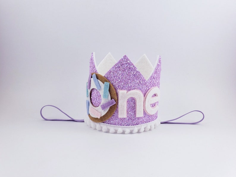 May include: Purple glitter crown with a white felt base and a felt donut with sprinkles. The crown has a white felt trim and says "one" in pink felt letters.