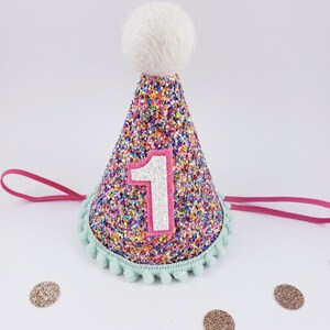 May include: A colorful glitter party hat with a white pom-pom on top and a pink felt number "1" on the front. The hat has a light blue trim with pom-poms around the bottom.