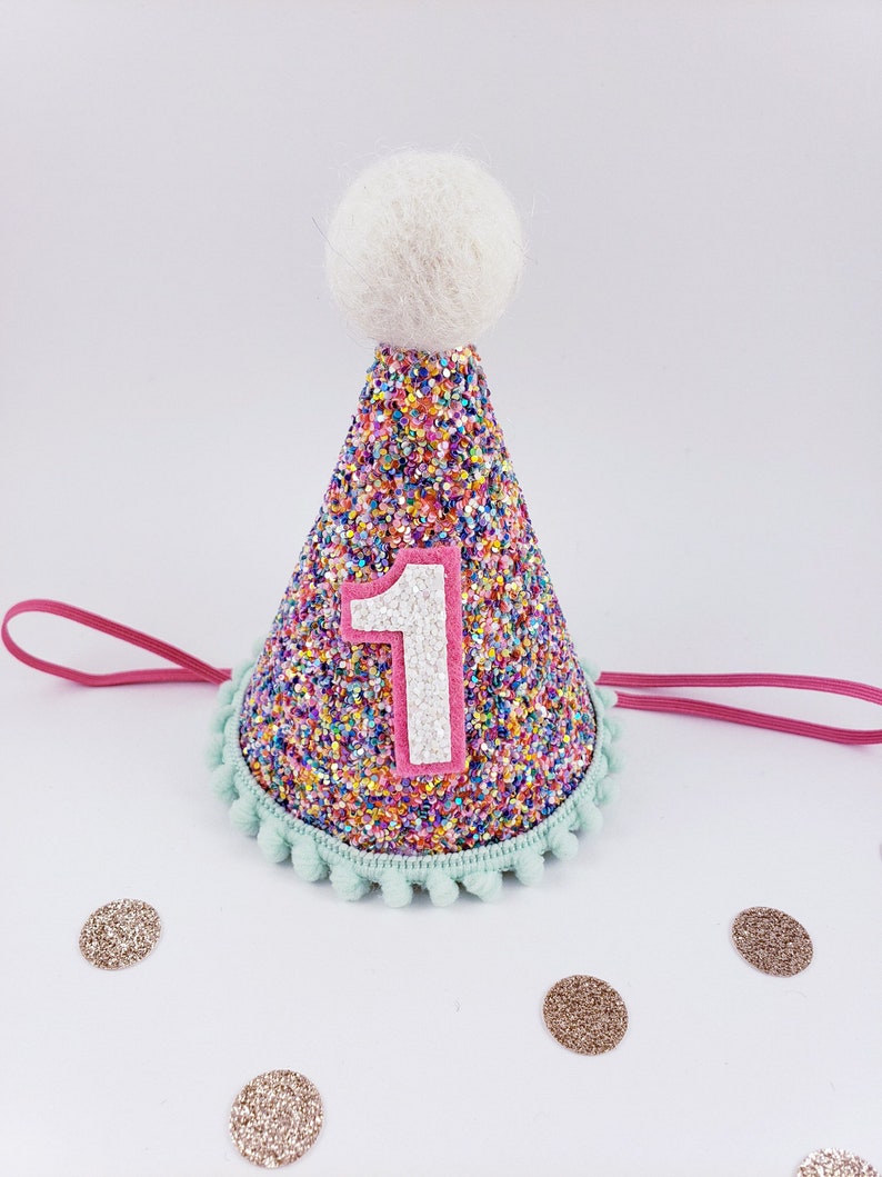 May include: A colorful glitter party hat with a white pom-pom on top and a pink felt number "1" on the front. The hat has a light blue trim with pom-poms around the bottom.