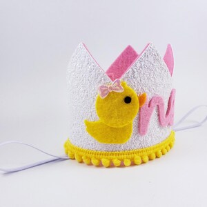 May include: White felt crown with pink and yellow felt accents. The crown features a yellow felt chick with a pink bow and the word "one" in pink felt letters. The crown has a white elastic band.