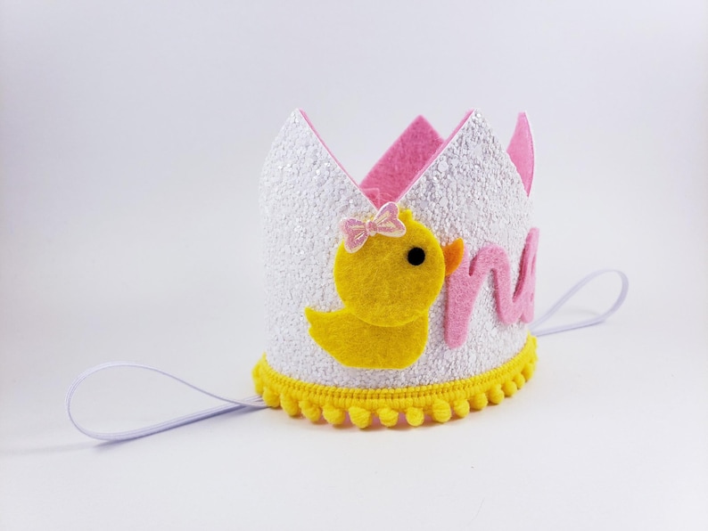 May include: White felt crown with pink and yellow felt accents. The crown features a yellow felt chick with a pink bow and the word "one" in pink felt letters. The crown has a white elastic band.