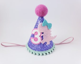 May include: Purple glitter party hat with a pink felt dinosaur, a blue bow, and a white felt number 3. The hat has a pink elastic band and a green pom-pom trim.