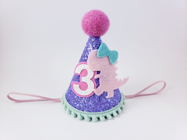 May include: Purple glitter party hat with a pink felt dinosaur, a blue bow, and a white felt number 3. The hat has a pink elastic band and a green pom-pom trim.
