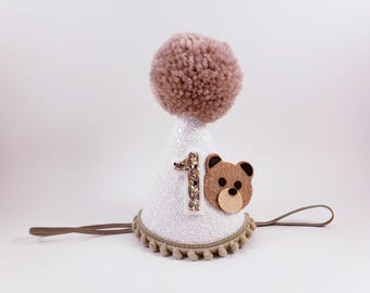 May include: A white, glitter-covered party hat with a brown pom-pom on top. The hat features a brown felt teddy bear face and the number "1" in gold glitter. A brown elastic band is attached.