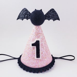 May include: Pink and black glitter party hat with a black bat on top and the number "1" on the front. The hat has a black elastic band.